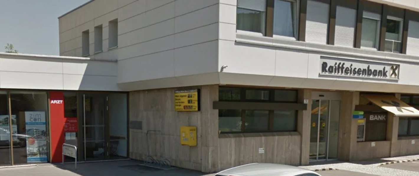 Eingang der Praxis mit rotem Schild „Arzt“ neben der Raiffeisenbank an der Hauptstraße